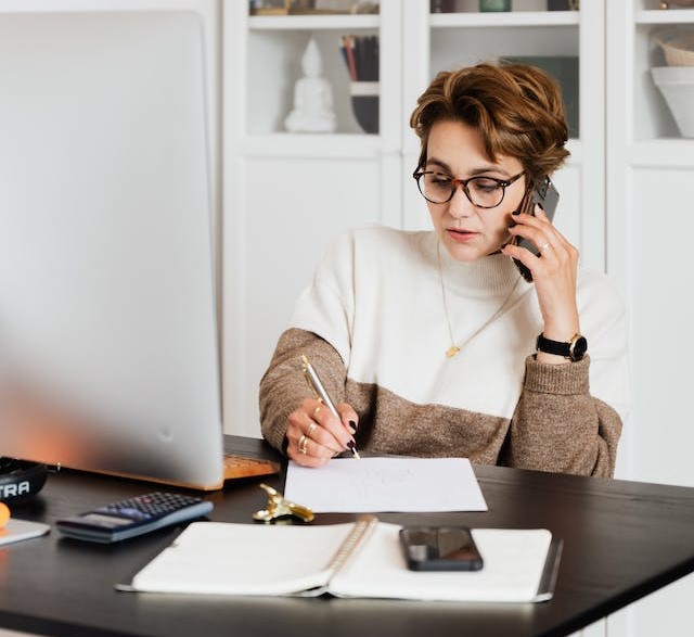 femme cheveux châtain et courts, au téléphone, porte un pull blanc et marron clair, des lunettes, sur un bureau noir. Derrière elle une bibliothèque blanche 